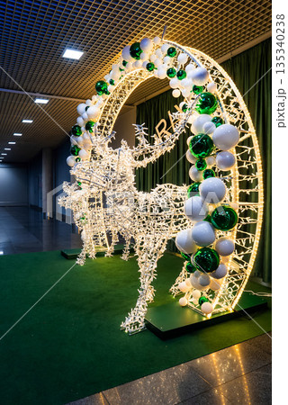 Two illuminated reindeer leap through a light wrapped arch in Riga, Latvia, on green turf with reflective tile flooring and a gridded ceiling, shot from a low angle. 135340238