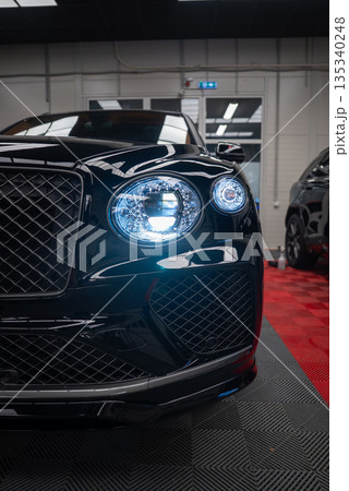 Close up three quarter view of a black Bentley Bentayga Speed indoors. Crystal cut LED headlamps glow, matrix grille and intakes show, red and black floor and white walls present. 135340248