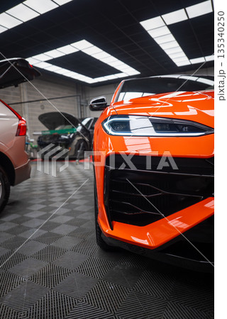 Close three quarter view shows orange Lamborghini Urus SE in a workshop, LED headlight, sculpted bumper, glossy black grille, cars nearby with hood and hatch open. 135340250