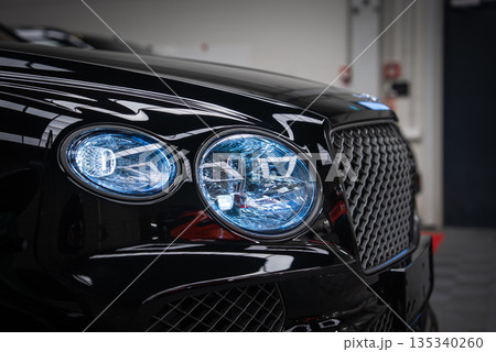 Close view of a black Bentley Bentayga Speed in a garage, twin crystal cut LED headlights with blue tinted illumination and diamond mesh grille with winged badge. 135340260