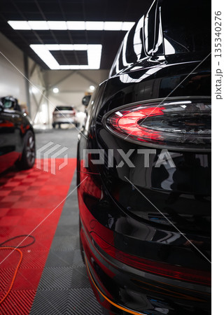 Glossy black Bentley Bentayga Speed rear close up shows oval taillamp, sculpted fascia, cool indoor lighting, red and gray floor mats, coiled power cable, parked cars. 135340276