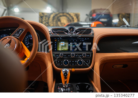 Bentley Bentayga Speed cockpit shows analog Breitling style clock, twin chrome vents, nav and FM screen, orange gear selector, and Bentley B emblem in indoor bay with soft lighting. 135340277