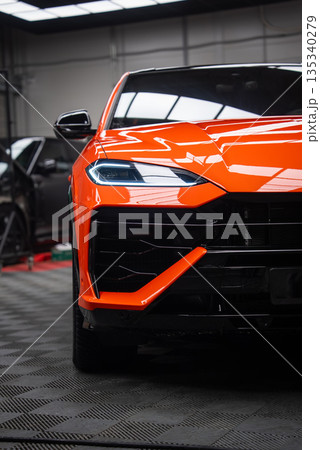 Close up shows orange Lamborghini Urus SE in an indoor workshop, with angular LED headlight, sculpted hood, black grille and orange aero blade under bright lights. 135340279