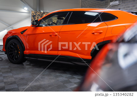 A close up side view shows an orange Lamborghini Urus SE parked in a modern workshop, with glossy paint, black wheels, orange calipers, checkered flooring, and detailing gear. 135340282