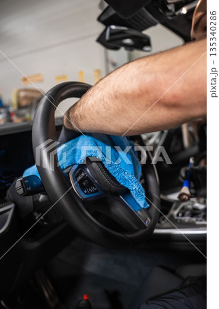 A person wipes a car steering wheel with a bright blue microfiber towel in a garage. Dashboard and controls are sharp, tools blur in back, soft indoor light shows care. 135340286