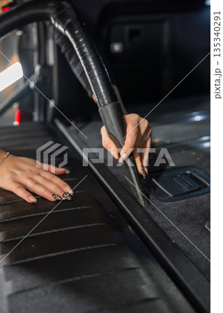A person vacuums the trunk seam of a car with a narrow crevice tool in a detailing bay, task lighting present, dark trim and leopard pattern nail art visible. 135340291