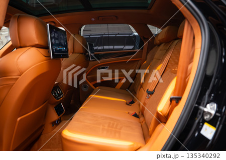 Close up rear cabin of Bentley Bentayga Speed shows quilted leather, B emblems on screens, rear entertainment tablets, wood inlays, panoramic glass roof, and climate controls. 135340292