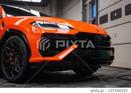 Close up low angle view shows the front left of an Orange Lamborghini Urus SE in an indoor service bay. Gloss black grille, LED headlight, Y accents, and orange calipers are visible. 135340293