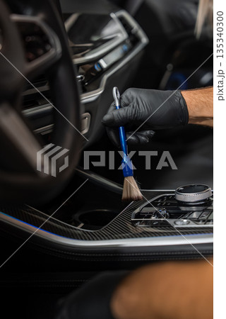 A gloved hand uses a detailing brush to clean dust around the gear selector and control dials on carbon fiber trim, inside a modern car with soft focused lighting. 135340300