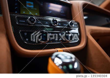 Close up indoor view shows carbon fiber trim, tan leather, knurled chrome dials, dual zone climate set to 22 C, Phone Nav and OK buttons, and a crystal like gear selector. 135340304
