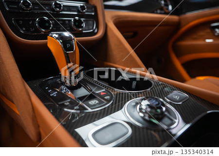 Close up interior shows crystal topped gear selector in Park, twin cupholders, carbon fiber trim, tan leather, knurled rotary controls, chrome buttons, and climate dials. Close up interior shows crystal topped gear selector in Park, twin cupholders, carbon fiber trim, tan leather, knurled rotary controls, chrome buttons, and climate dials. 135340314
