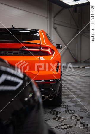 Three quarter rear view shows orange Lamborghini Urus SE in a workshop with angular LED taillight, quad exhaust tips, checkered floor, and overhead fluorescent lights. 135340320