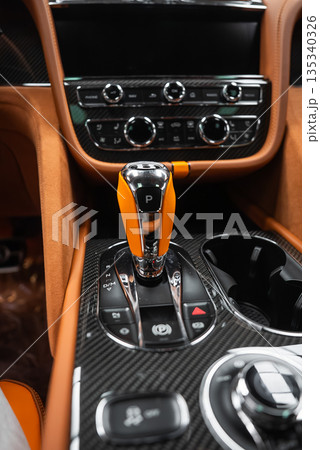 Close up indoor view of a Bentley Bentayga Speed center console, crystal and metal gear selector with P indicator, carbon fiber trim, tan leather, knobs, switches, cup holders. Close up indoor view of a Bentley Bentayga Speed center console, crystal and metal gear selector with P indicator, carbon fiber trim, tan leather, knobs, switches, cup holders. 135340326
