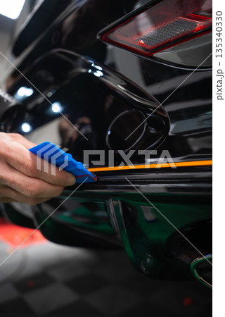 A car technician uses a blue squeegee to align an orange vinyl stripe on a glossy black sports car rear bumper in a workshop, tail light and dual exhaust visible. A car technician uses a blue squeegee to align an orange vinyl stripe on a glossy black sports car rear bumper in a workshop, tail light and dual exhaust visible. 135340330