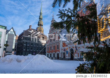 Fresh snow piles in Riga, Latvia Old Town square as lights glow on a Christmas tree. House of the Blackheads and St. Peter's Church spire rise in soft daylight. 135340332