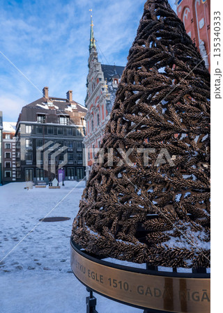 A bronze fir branch sculpture with snow and a ring marked 1510 stands in Riga Old Town, with House of the Blackheads and gabled facades under a crisp sky. A bronze fir branch sculpture with snow and a ring marked 1510 stands in Riga Old Town, with House of the Blackheads and gabled facades under a crisp sky. 135340333