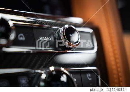 Close interior view shows a knurled chrome radio knob with arrow icon and RADIO label, carbon fiber, polished metal, and tan leather, in soft shallow depth lighting. Close interior view shows a knurled chrome radio knob with arrow icon and RADIO label, carbon fiber, polished metal, and tan leather, in soft shallow depth lighting. 135340334