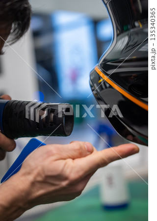 A car worker uses a heat gun to apply vinyl wrap on a glossy black panel with an orange stripe. Indoor workshop, green floor mat, blurred tools, shallow depth of field. 135340365