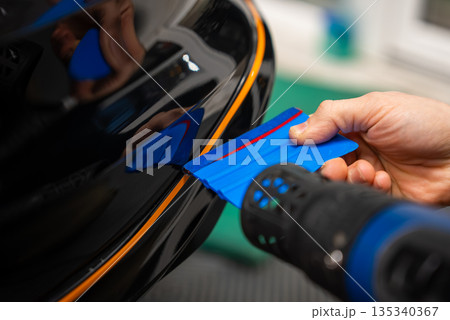 A car technician uses a blue squeegee and a heat gun to fit an orange pinstripe on a glossy black bumper edge in an indoor auto wrap workshop, emphasizing precision. 135340367