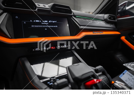 Close interior view of an Orange Lamborghini Urus SE in a workshop. Hexagonal vents, angular switchgear, Lamborghini script, and Russian prompt on screen are visible. Close interior view of an Orange Lamborghini Urus SE in a workshop. Hexagonal vents, angular switchgear, Lamborghini script, and Russian prompt on screen are visible. 135340371
