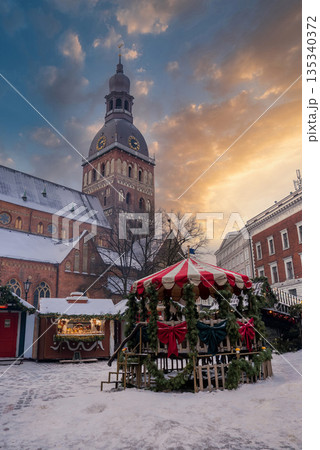 Snowy Christmas market in Riga, Latvia, shows carousel, wooden stalls, garlands, and the Riga Cathedral clock tower at dusk as locals and visitors gather. Snowy Christmas market in Riga, Latvia, shows carousel, wooden stalls, garlands, and the Riga Cathedral clock tower at dusk as locals and visitors gather. 135340372