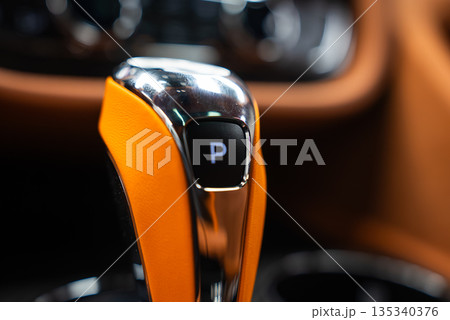 Close up shows Bentley Bentayga Speed gear selector with chrome and orange leather, illuminated P park indicator, tan upholstery, and blurred metallic switchgear. Close up shows Bentley Bentayga Speed gear selector with chrome and orange leather, illuminated P park indicator, tan upholstery, and blurred metallic switchgear. 135340376