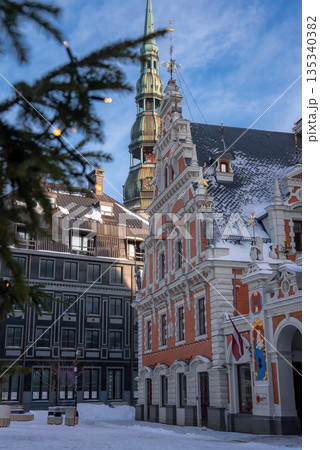 House of the Blackheads stands in Riga, Latvia as snow dusts cobblestones. St. Peter's Church spire rises behind. A Latvian flag and fairy lights signal Christmas afternoon. 135340382