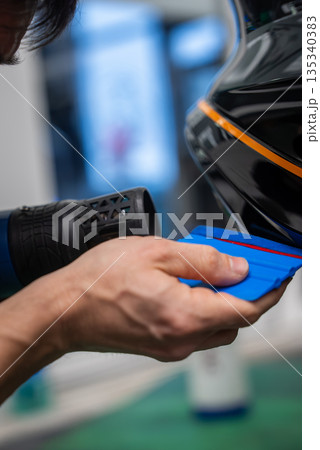 Close up shows a worker applying vinyl wrap to a glossy black panel using a heat gun and blue squeegee, forming around an edge with an orange stripe in a workshop. Close up shows a worker applying vinyl wrap to a glossy black panel using a heat gun and blue squeegee, forming around an edge with an orange stripe in a workshop. 135340383
