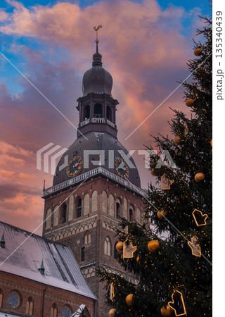 Riga Cathedral dome and clock tower rise at winter sunset in Riga, Latvia. A lit Christmas tree with gingerbread lights and baubles stands by snow rooftops. Riga Cathedral dome and clock tower rise at winter sunset in Riga, Latvia. A lit Christmas tree with gingerbread lights and baubles stands by snow rooftops. 135340439