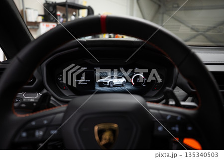 Close up from the driver seat shows the Lamborghini crest wheel and red marker, with the Urus digital cluster active, in a workshop under soft indoor lighting. 135340503