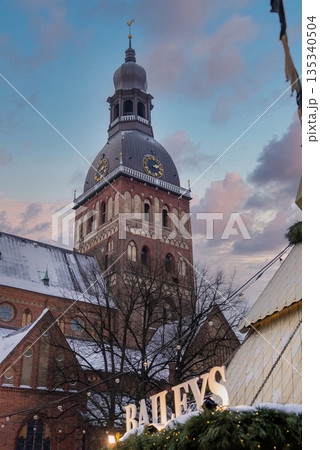 Riga Cathedral, or Rigas Doms, rises over Old Town in Latvia as festive lights, evergreen garlands, and a BAILEYS sign glow at twilight with snow on the spire. 135340504