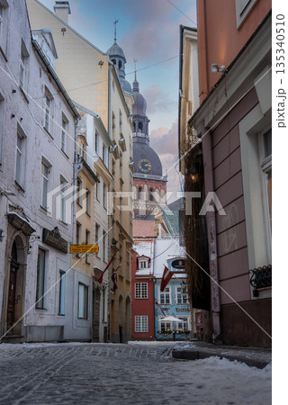 A narrow cobblestone lane in Riga Old Town leads to Riga Cathedral. Snow rests on edges and sills. Latvian flags and signs appear. Soft cool daylight sets a calm mood. 135340510