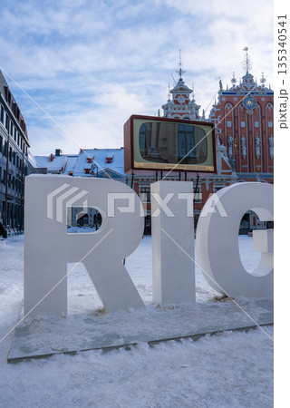 Snow covers Riga Old Town as the white RIGA sign stands with a retro TV frame. The House of the Blackheads rises behind. Soft daylight and blue sky suggest Christmas. Snow covers Riga Old Town as the white RIGA sign stands with a retro TV frame. The House of the Blackheads rises behind. Soft daylight and blue sky suggest Christmas. 135340541