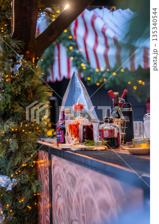 A festive stall in Riga, Latvia shows mulled wine, syrups, and spices beside an ice cone, under a red and white canopy with garlands and warm string lights at evening. 135340544