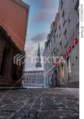 Narrow cobblestone alley in Riga Old Town opens to a square, pastel facades, neon VOLMAR sign, and St. Peter's Church spire in soft winter daylight with light snow. 135340545