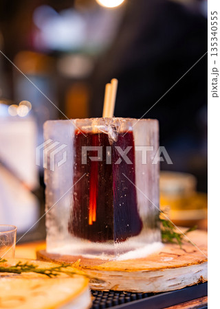 A steaming mulled wine in a clear ice block sits on a birch coaster in Riga, Latvia. Warm market lights blur behind, with sprigs of greenery suggesting festive winter mood. A steaming mulled wine in a clear ice block sits on a birch coaster in Riga, Latvia. Warm market lights blur behind, with sprigs of greenery suggesting festive winter mood. 135340555