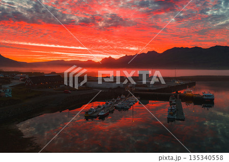 A small Icelandic harbor during a red and orange sunset, with docked boats, calm reflective water, rugged mountain silhouettes, and nearby buildings. 135340558