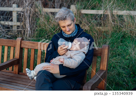 A young father bottle feeding his baby while walking outdoors. Feeding the baby. Dad smiling, spending time with his little son in the park. Family time together. Everyday parenting lifestyle. 135340939