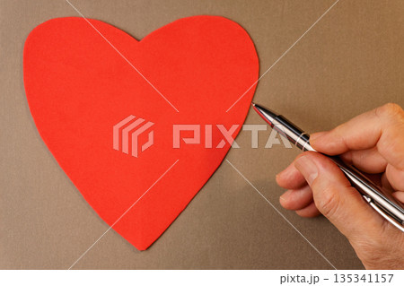 Man hand writing on blank red heart card with pen on brown background. Man hand writing on blank red heart card with pen on brown background. 135341157