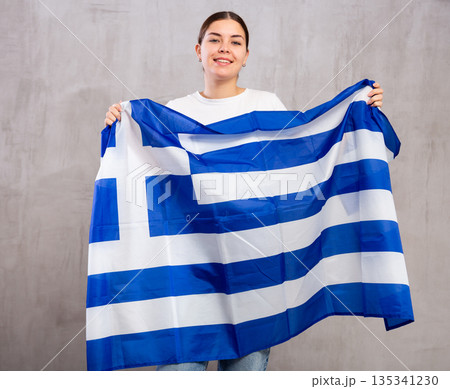 Joyous young woman holding Greece flag against unicoloured background Joyous young woman holding Greece flag against unicoloured background 135341230