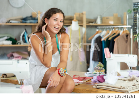 Carefree young female tailor sitting on workbench in workshop Carefree young female tailor sitting on workbench in workshop 135341261