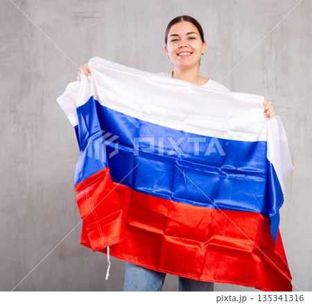 Smiling young woman standing with flag of Russia against gray wall background 135341316