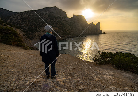 Woman watching sunset over rugged seascape mountains feeling peaceful solitude 135341488