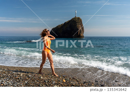 Woman beach cross. Young lady walks on pebble shoreline near iconic cape rock on a sunny summer day. 135341642