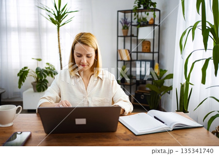 Focused woman working on laptop in cozy home office Focused woman working on laptop in cozy home office 135341740