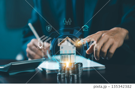 A hand pointing to a miniature wooden house model on stacked coins, surrounded by digital icons representing family, security, property, and wealth management/investment. A hand pointing to a miniature wooden house model on stacked coins, surrounded by digital icons representing family, security, property, and wealth management/investment. 135342382