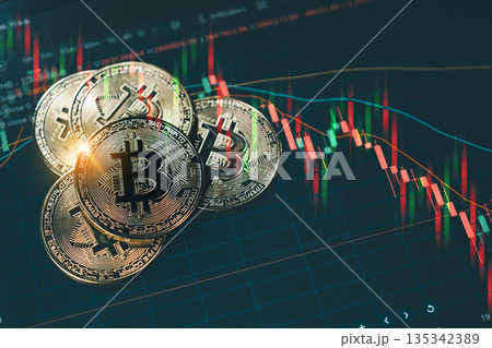 A shiny gold Bitcoin coin stacked on other coins, set against a dark background with red and green candlestick charts illustrating a volatile cryptocurrency market downtrend. 135342389
