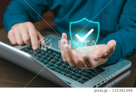 A close-up of a person's hand holding a glowing blue security shield icon with a checkmark above a laptop keyboard, representing data protection and cyber security. 135342394
