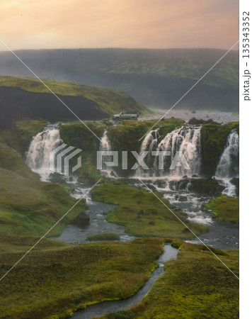 A small, multi tiered waterfall flows over moss covered rocks in a lush Icelandic setting. A rugged vehicle sits nearby, surrounded by streams and hills. 135343352
