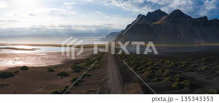 Aerial view of Stokksnes Peninsula in Iceland, showing a dirt road on black sand, Vestrahorn Mountain, green mounds, and a reflective water body. 135343353